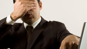 Man with his eyes covered pointing at a computer. 